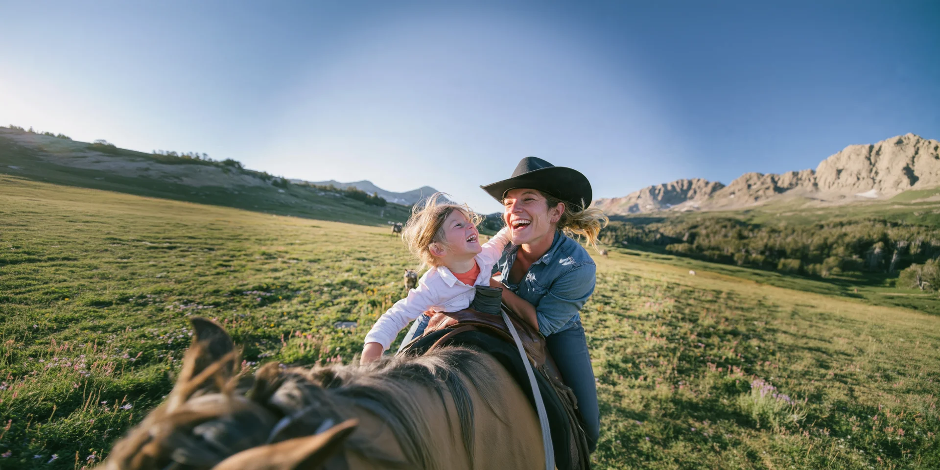A mother and daughter laughing together on horseback in an open mountain meadow